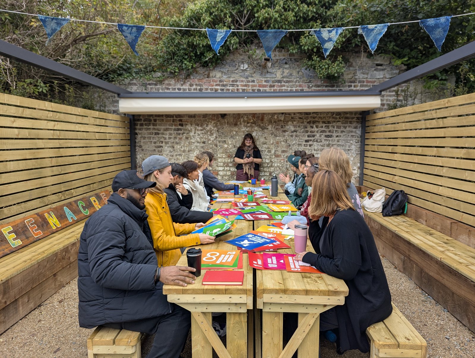 A class seated along a long wooden table in a covered outdoor
               classroom at Greenacre, working with Sustainable Development Goal
               cards, with the lecturer standing at the far end