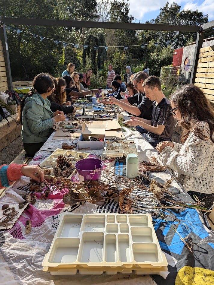 Students seated along a long outdoor table under bunting, sorting
               foraged pine cones, seed heads and dried plants into trays