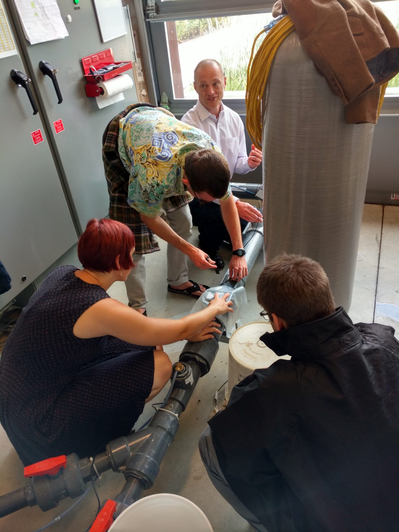 Students and a founder crouched around exposed pipework in a plant
               room, fitting a sensor to a water main